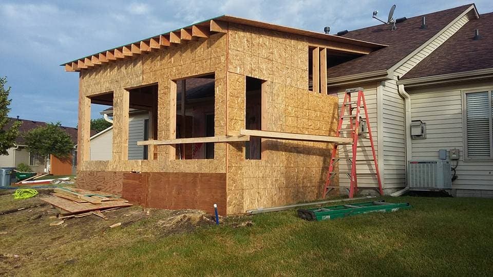 An addition framed and sheathed against the side of an existing house, rafters on and window openings cut.