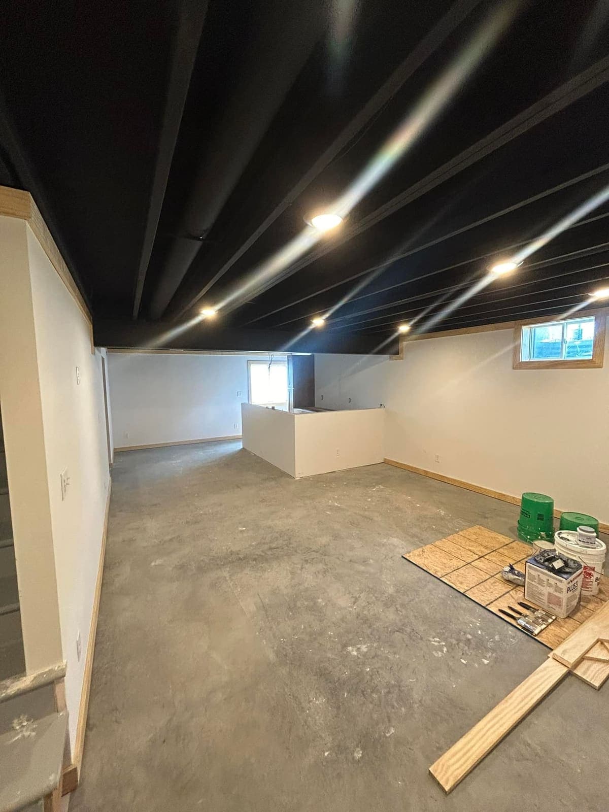 A basement with the joists painted out black and recessed lights in, standing on a bare concrete floor.