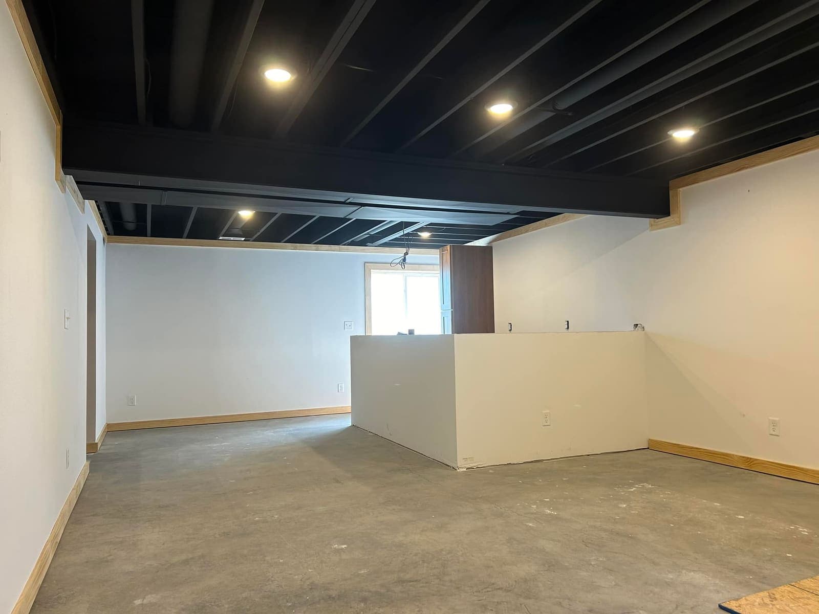 A basement painted and lit, with a finished half wall and the concrete floor waiting on its covering.