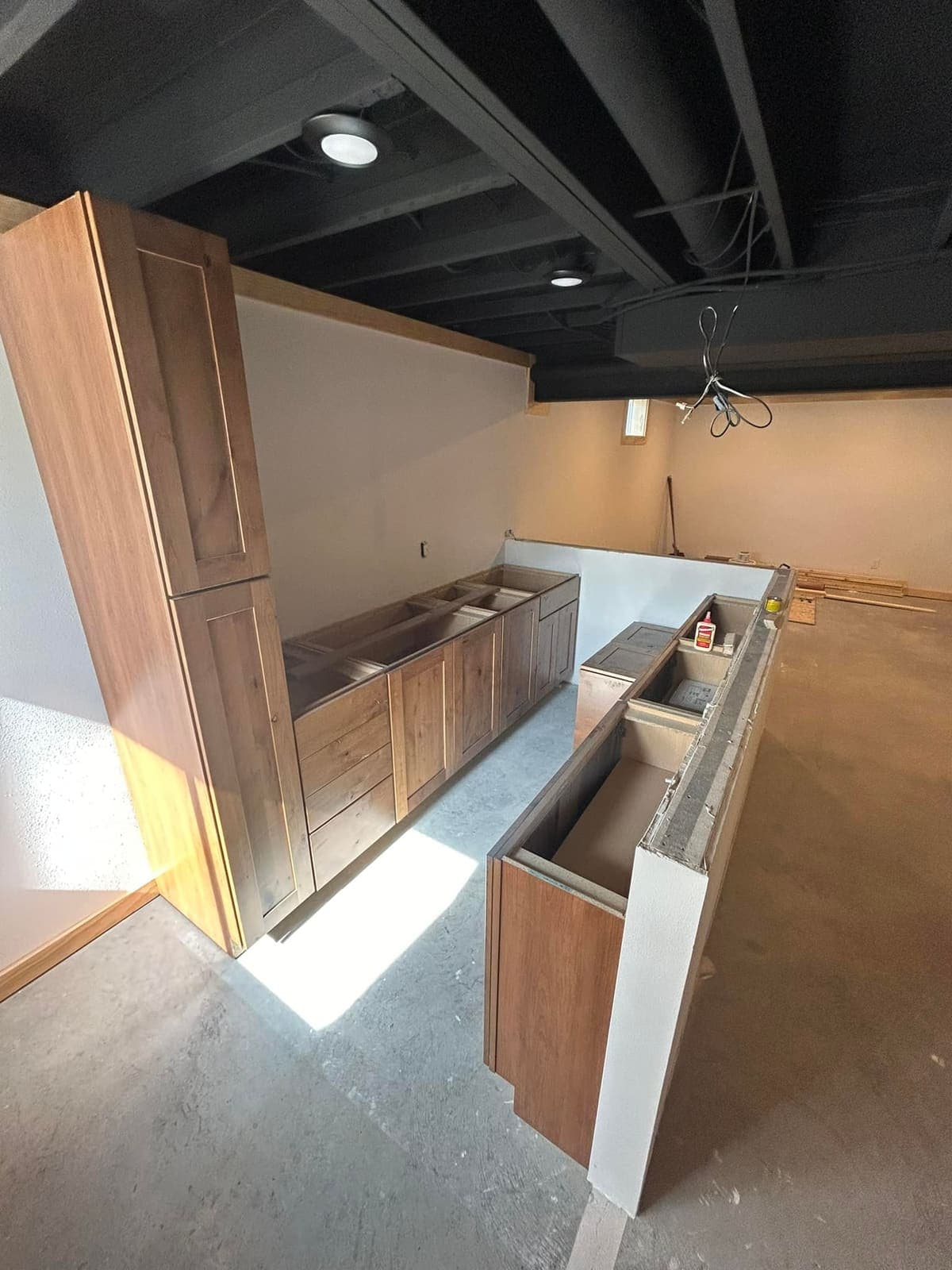 Bar cabinetry being set into a basement under a painted ceiling and pendant rough-ins.