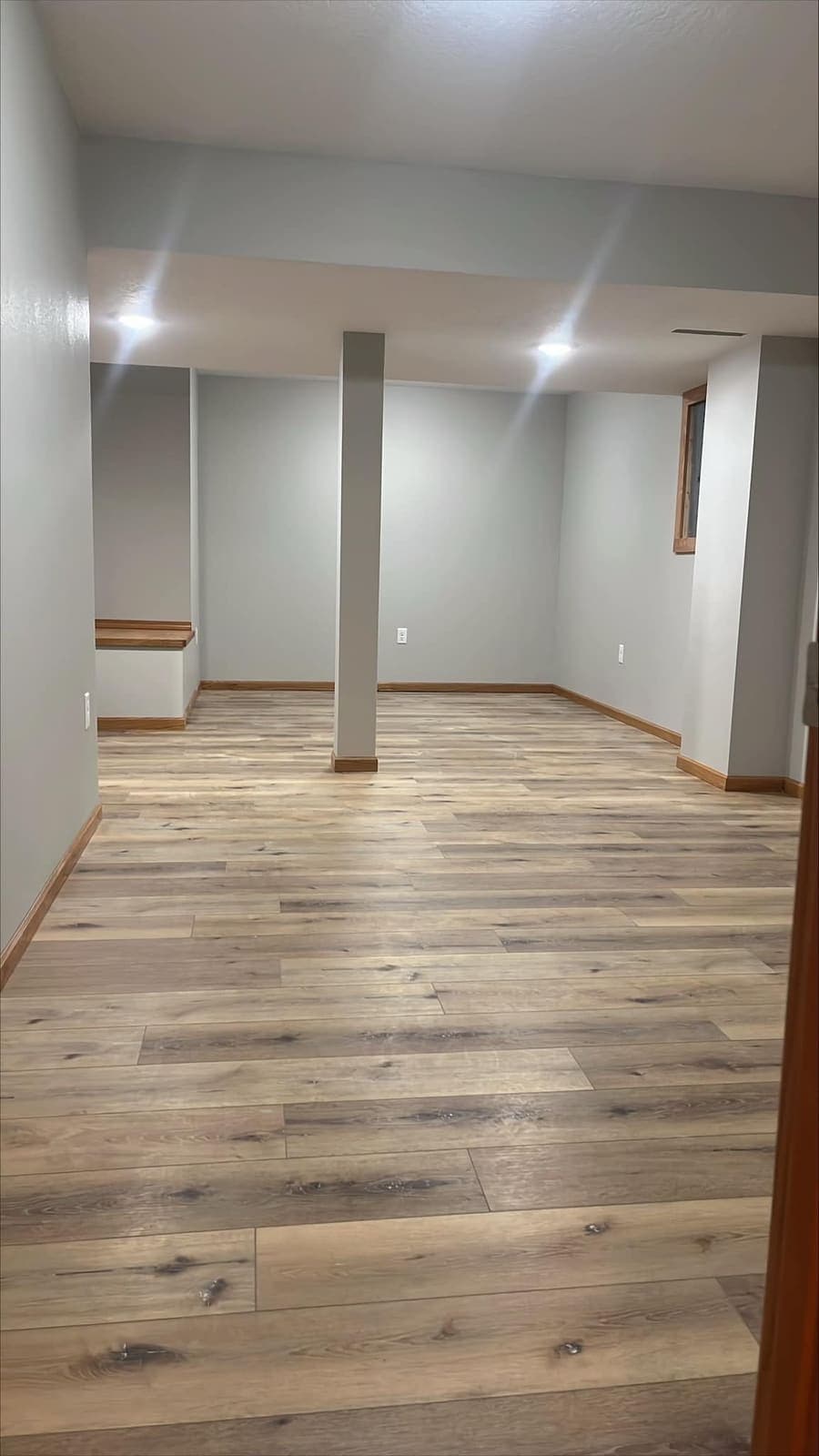 A finished basement room with plank flooring laid through it and a window in the far corner.