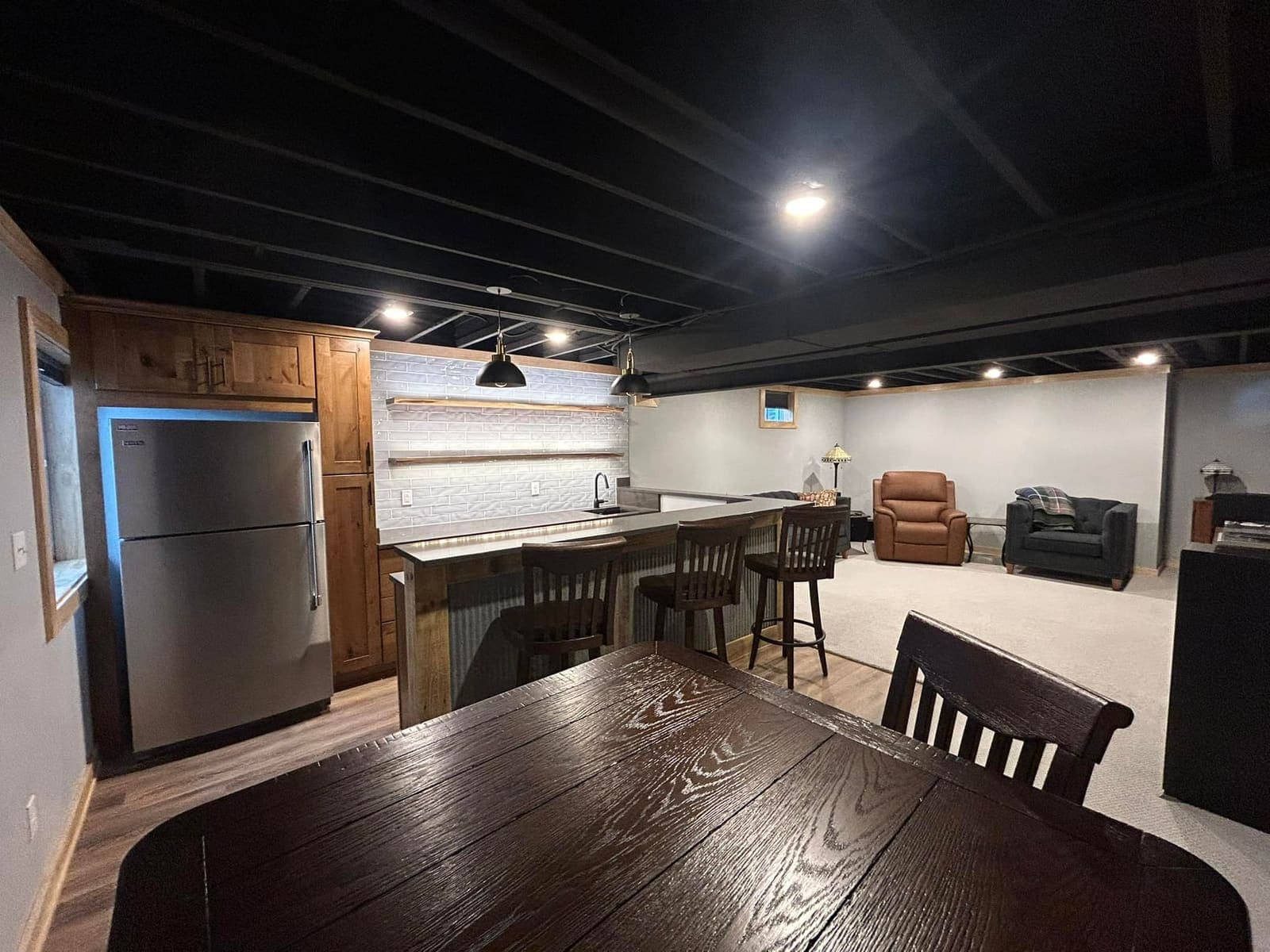 A finished basement bar with counter stools, a refrigerator and a dining table in the foreground.