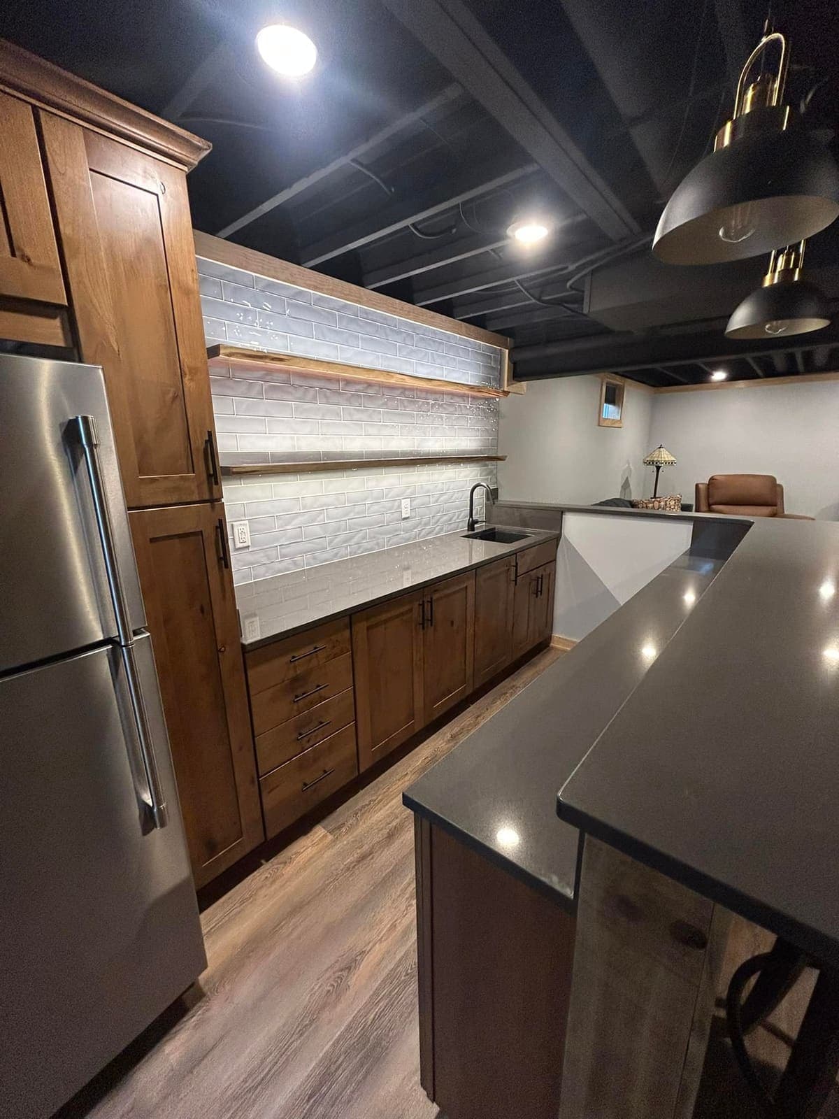 A basement kitchenette with stained cabinetry, a stone counter and a tiled wall behind the sink.
