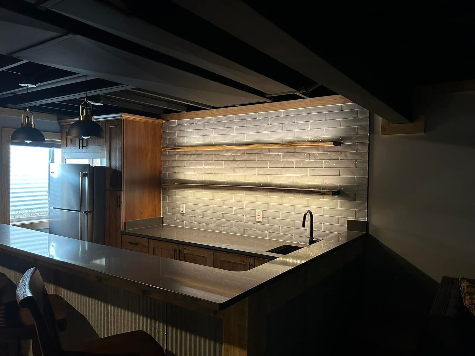 A basement bar with a tiled backsplash, lit floating shelves and a corrugated metal counter front.