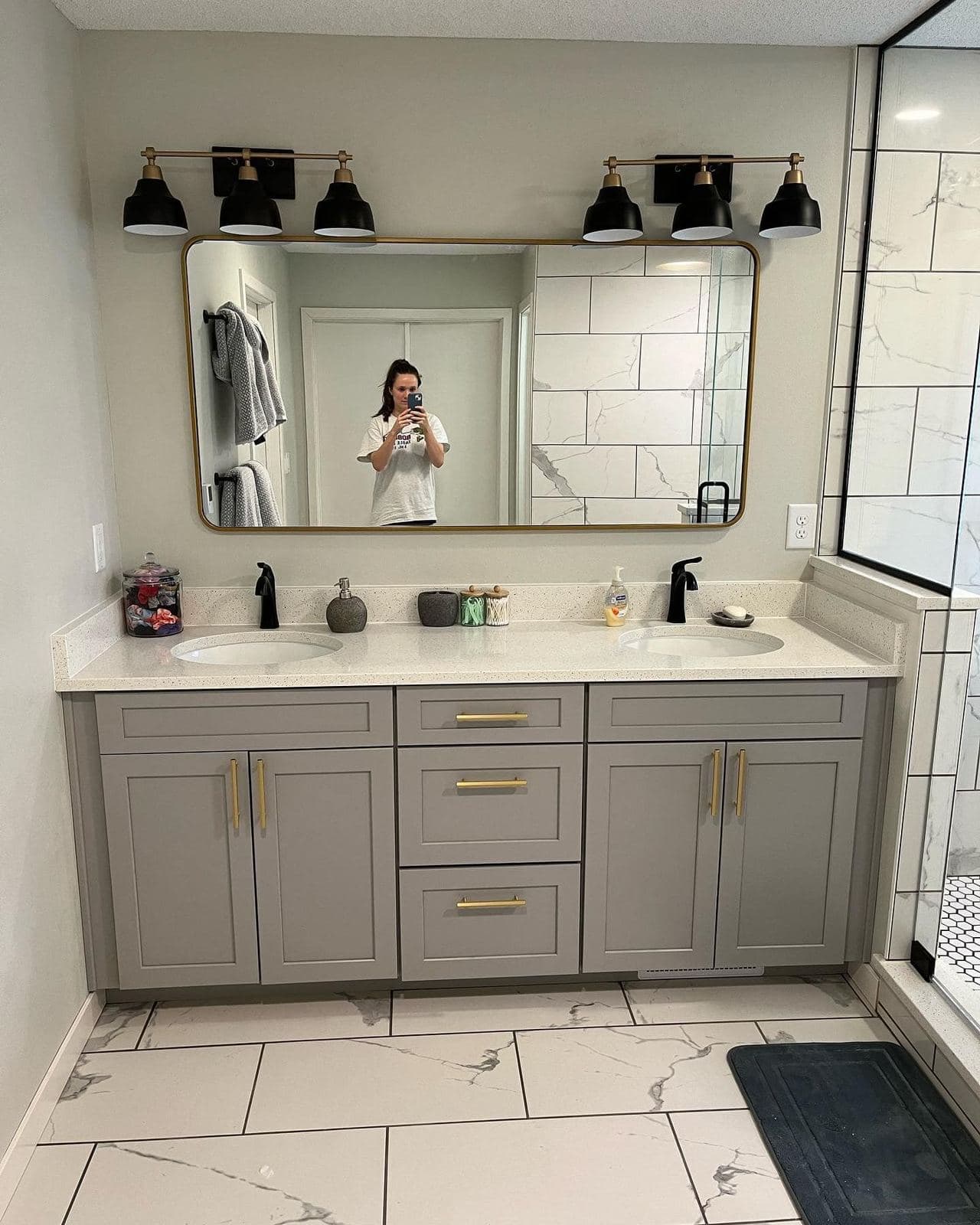 A double vanity with two rectangular sinks, gold hardware and black sconces flanking a large framed mirror, over marble-look tile.
