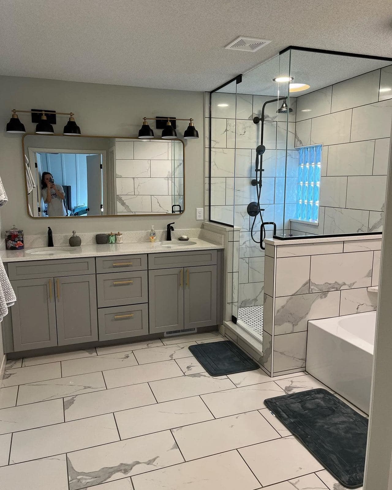 A remodeled bathroom with a glass shower, marble-look wall tile and a long vanity with black fixtures.