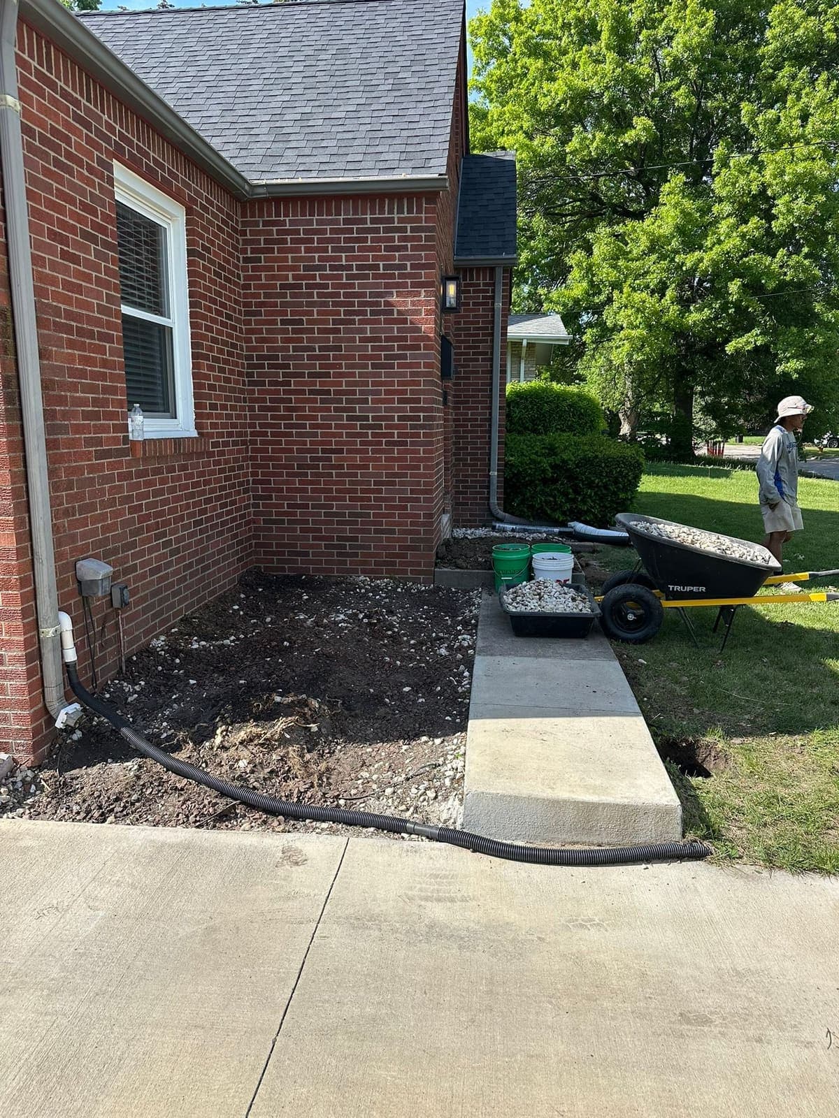 A new concrete sidewalk placed alongside a brick house, with the ground either side still open.