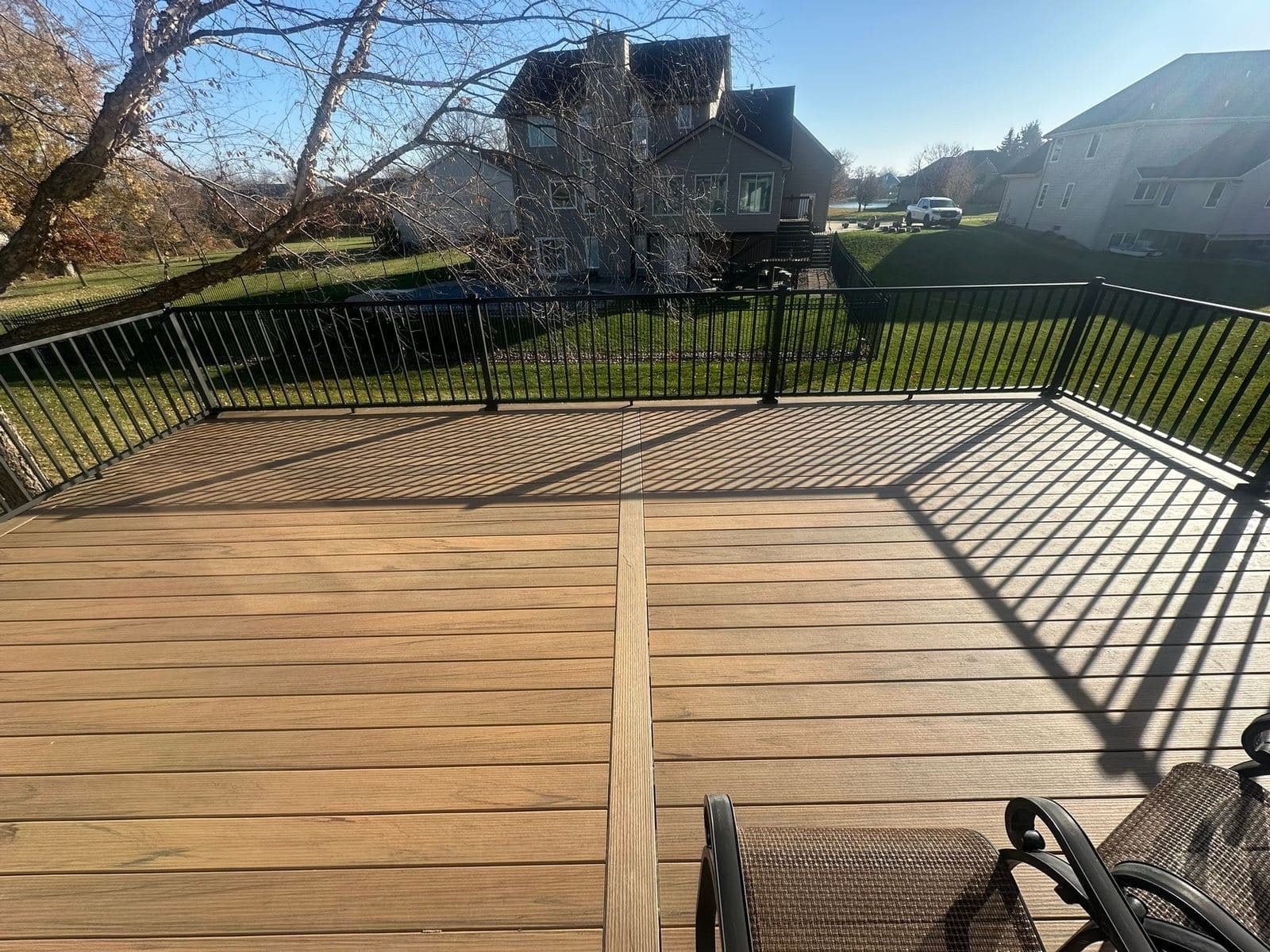 A large stained deck with black railing photographed in autumn, with mature trees and neighboring homes beyond.