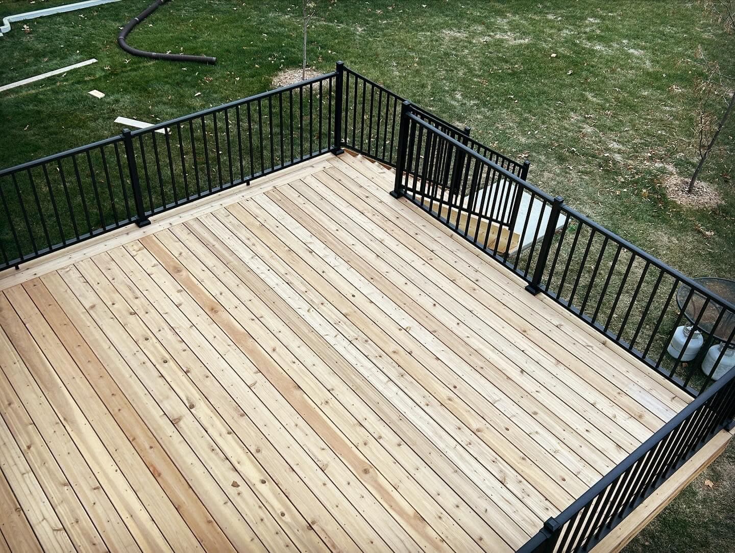 A newly built cedar deck seen from above, framed on three sides by black aluminum railing over a green lawn.