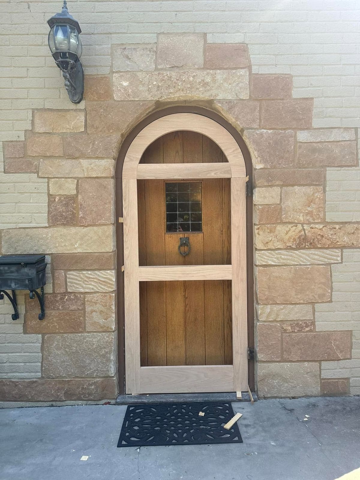 A custom arched wood door with a leaded glass insert, set into a cut stone surround beneath a lantern sconce.