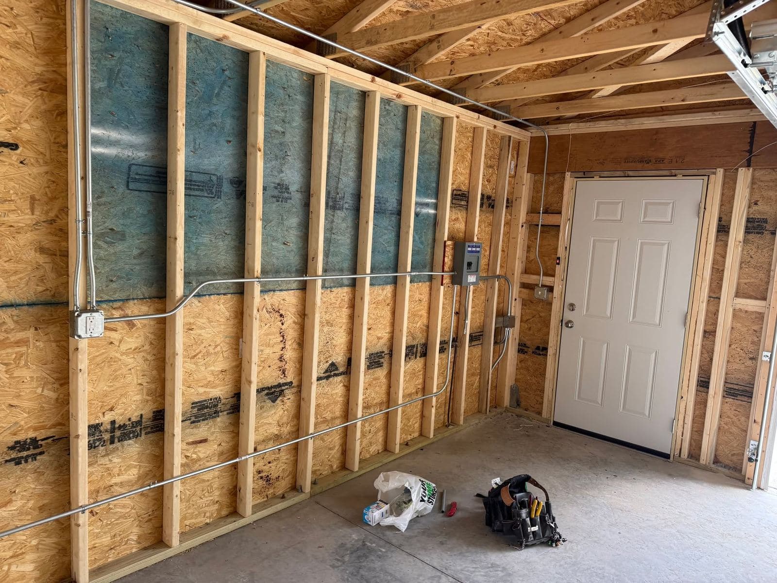 Electrical rough-in along a garage stud wall with conduit, junction boxes and a finished service door.