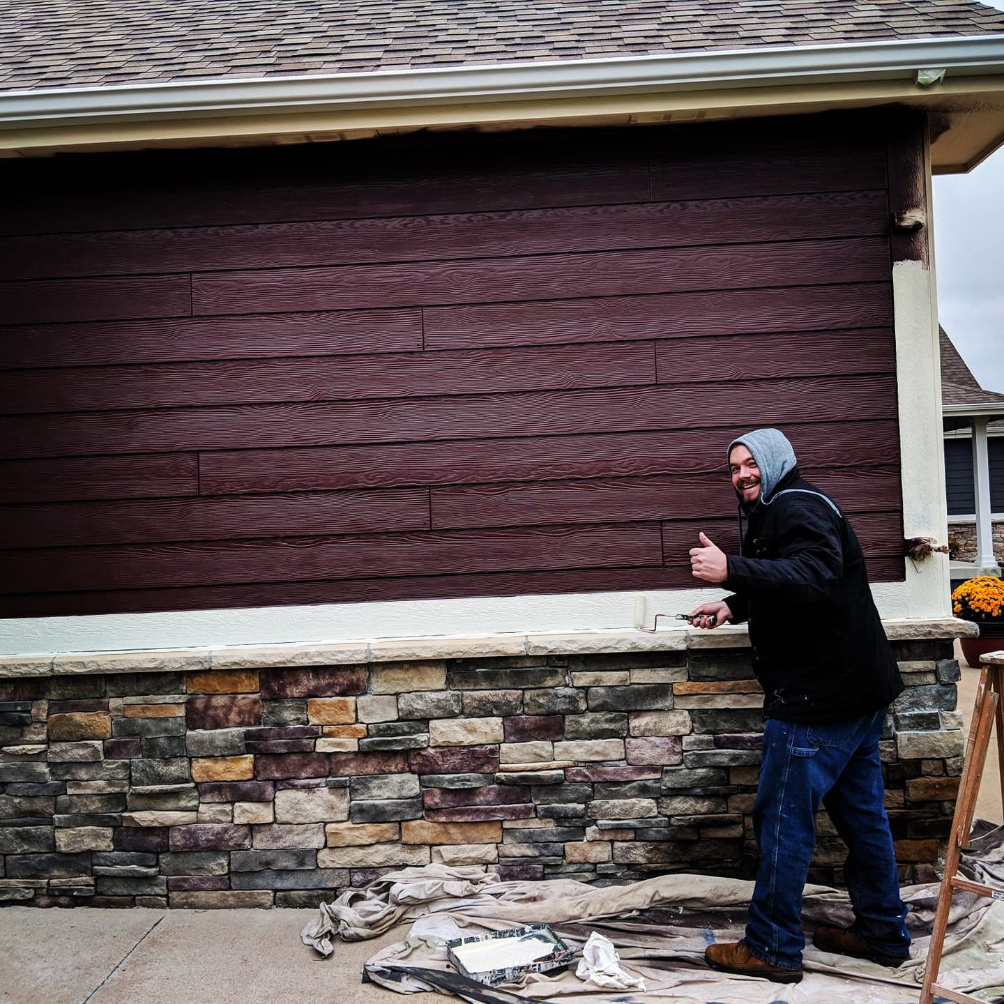 A painter cutting in fresh deep red siding paint above a stone veneer base, with drop cloths laid out below.