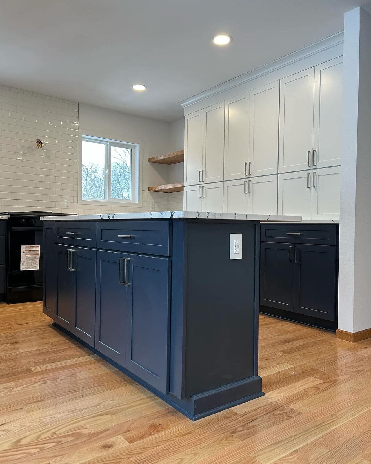 A kitchen with a deep navy island, white perimeter cabinetry and wood open shelving beside the range wall.