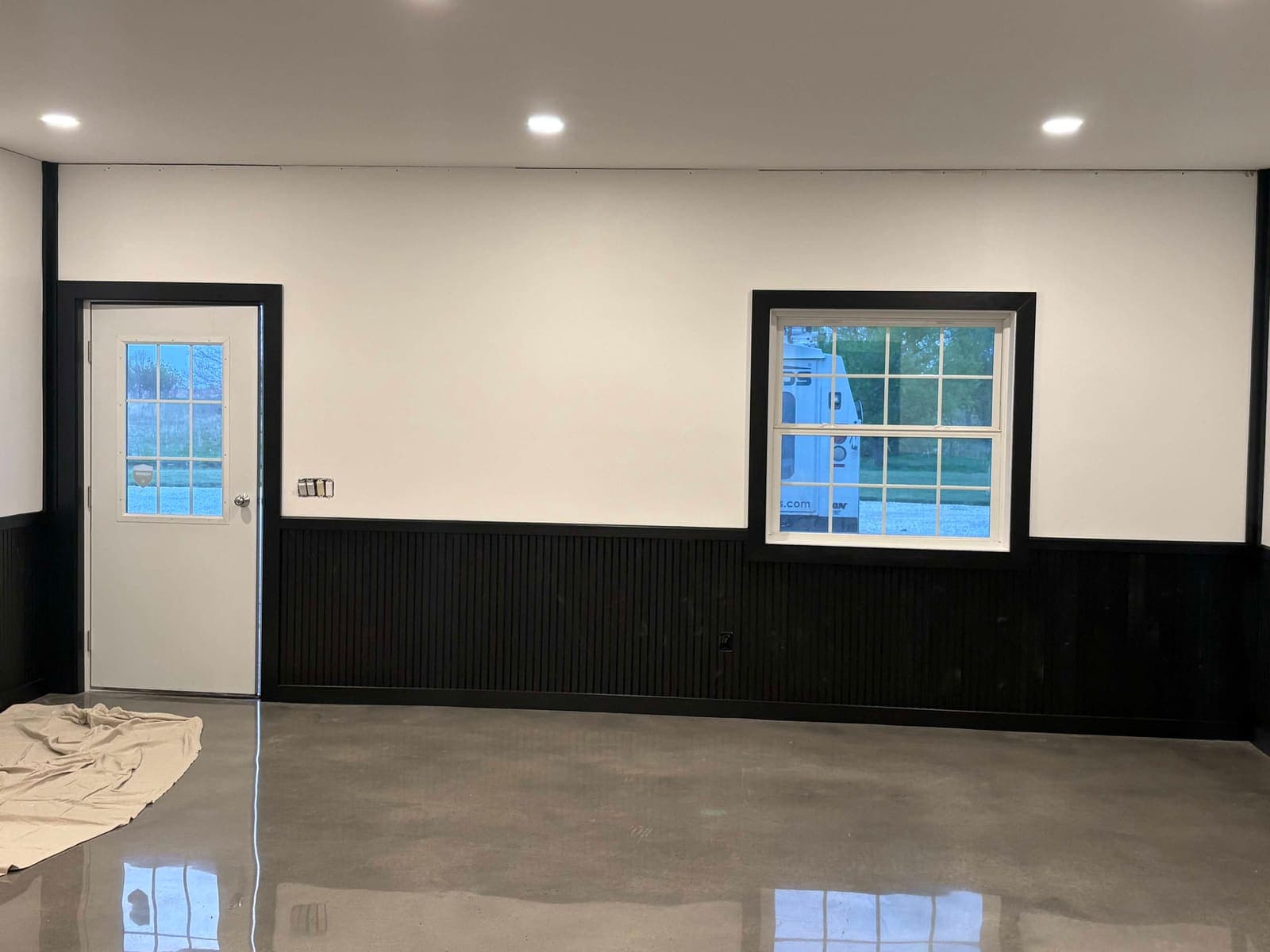 A room painted white above black wainscot, with black-trimmed door and window and a drop cloth still down.