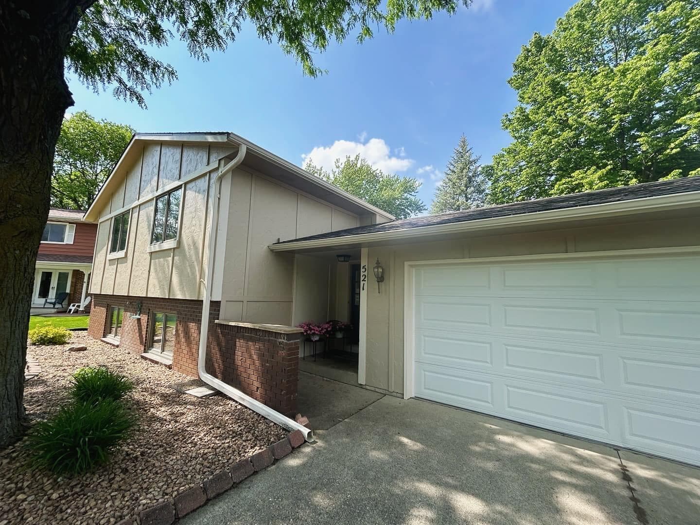 A tan-sided home and attached garage with white trim, shot from the driveway under mature trees.